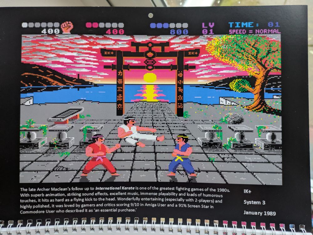 A screenshot of the Amiga conversion of the C64 game IK+. It is almost too difficult to describe it's beauty. A square-laved grey courtyard leads up to a blue lake, or the sea, with the sun setting behind it casting rippling reflections of pink and yellow on the water surface. A Japanese-style pergola structure frames this at the back of the courtyard, a tree stands off back right and brown black low mountains frame the lake, with a pink sky dotted with white clouds. Prominent at the front of the courtyard, three people are doing battle by karate. There is a red clad, white and blue clad combatant. In the middle, the white (the player) combatant is in the air, mid-way through a move called flying kick. In a ready defensive stance, the red and blue combatants stand either side, facing the white combatant. 