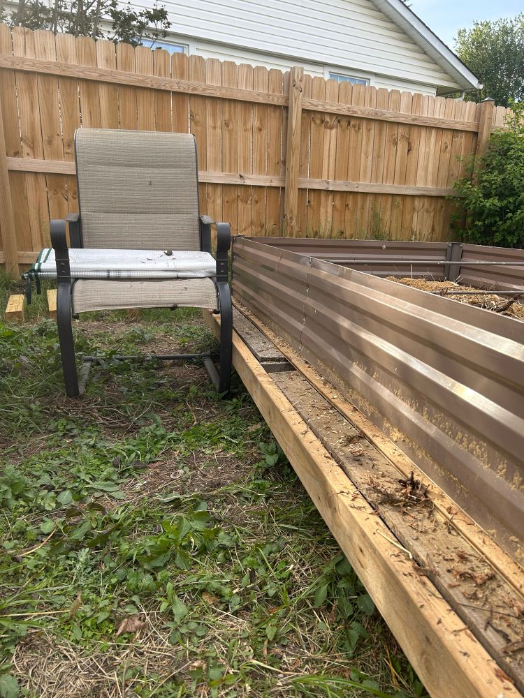 The base of the greenhouse with a yard chair next to it showing that the wooden frame it is sitting on puts it at a good relative height for the chair