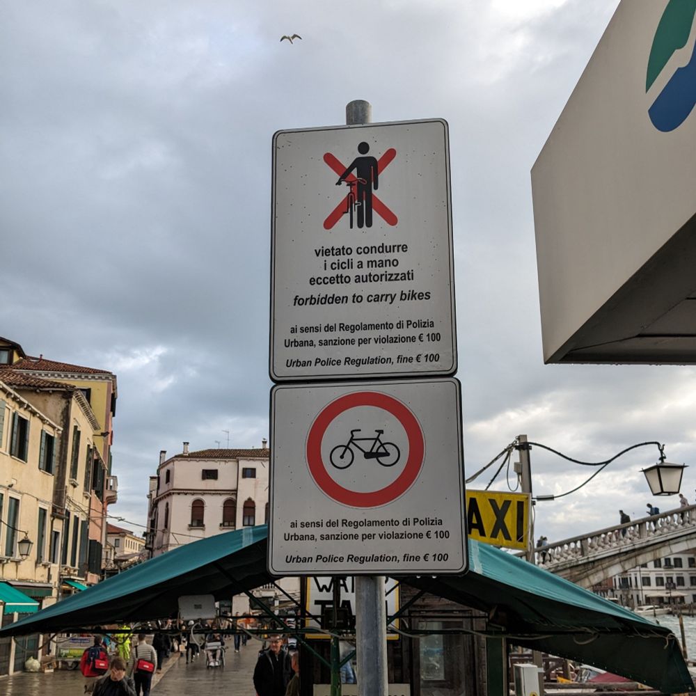 Ein Blechschild, beschriftet auf Italienisch, verbieten das Fahren mit dem Fahrrad. Darüber ein weiteres Schild, welches auch noch das mit-sich-führen eines Fahrrads unter Strafe stellt.