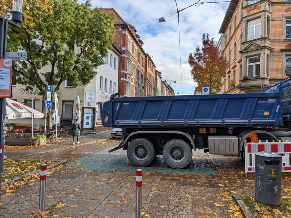 Ein Kieslaster, abgestellt quer über die Straße. Es bleibt nur ein schmaler Durchlass. Unter dem Laster vage erkennbar das Türkis der Fahrradstraße.