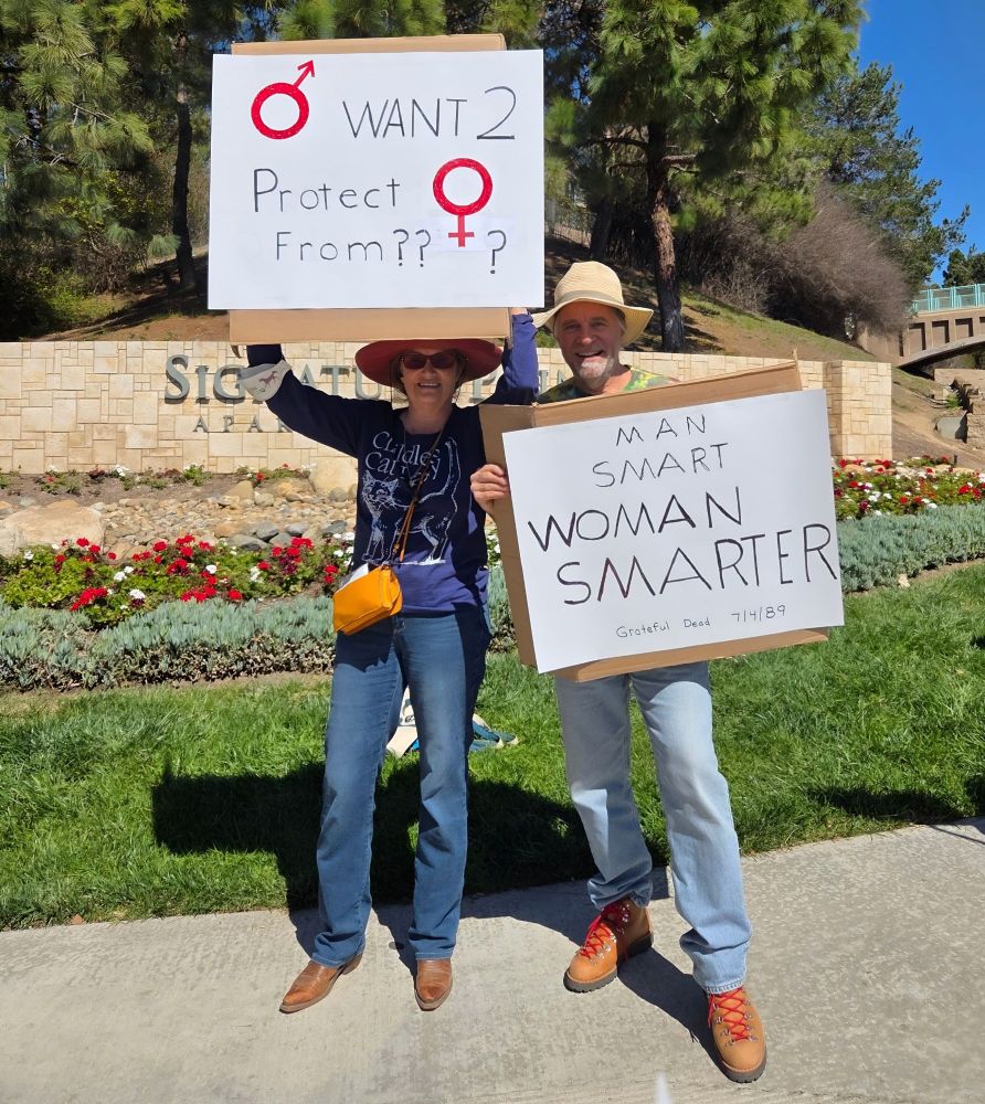 Woman holding a sign with (symbol for man) followed by want to protect (symbol for woman) from ??? Man holding sign saying 'man smart woman smarter' with Grateful Dead song reference