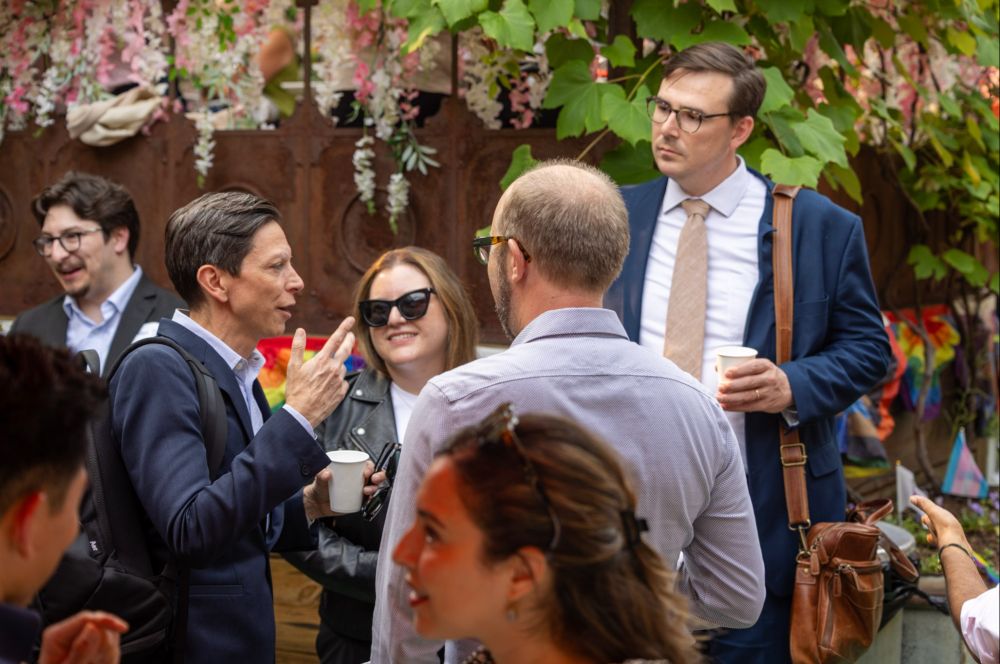 Five attendees chat in a circle at the center of the image, with several others in the foreground and background. 