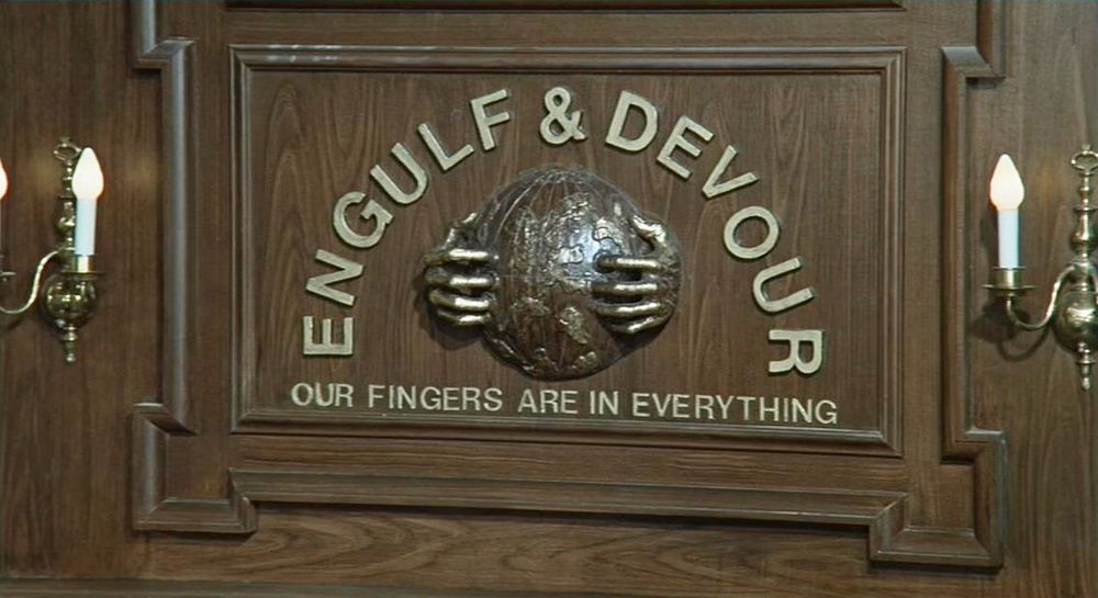 still from Mel Brook's "Silent Movie" of the logo for the evil corporation Engulf & Devour. It shows two hands clutching the world with the name   Engulf & Devour arched over it, and underneath it says "OUR FINGERS ARE IN EVERYTHING"