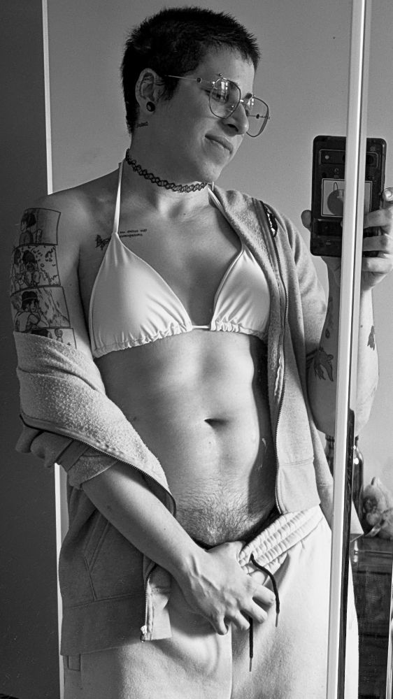 A selfie of Sable in the mirror. She's holding her phone in one hand, and pulling her pants down with the other, showing her bush a little. This time she's smiling at the phone.