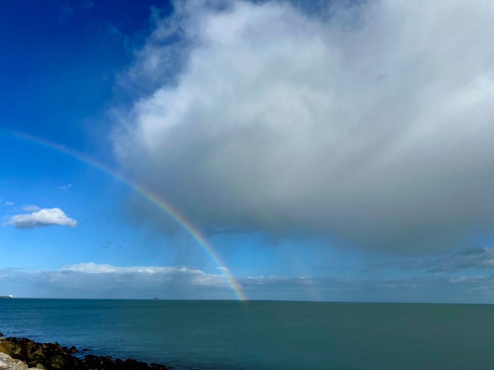 Seascape with massive cloud that is shedding rain, blue sky behind it and rainbow in front. It’s all happening! 