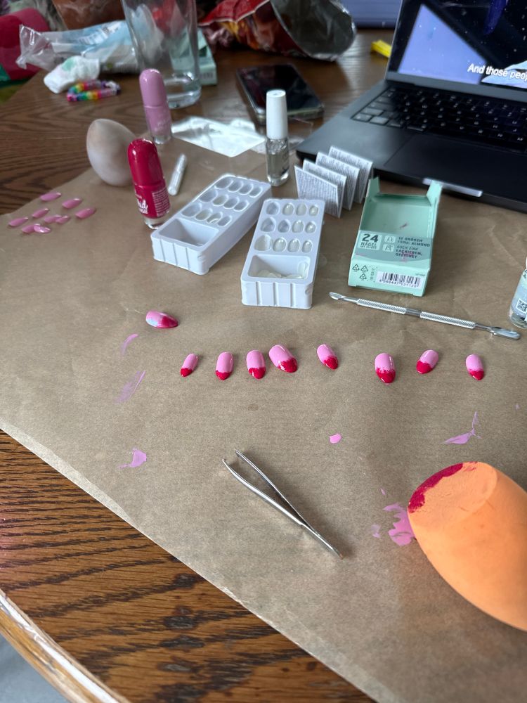A spread of press on nails that have been painted pink with red tips  they’re on top of a baking sheet covered in splotches of nail polish with makeup sponges covered in matching red nail polish. Towards the back are the various bottle of polish and top coat next to a laptop. 