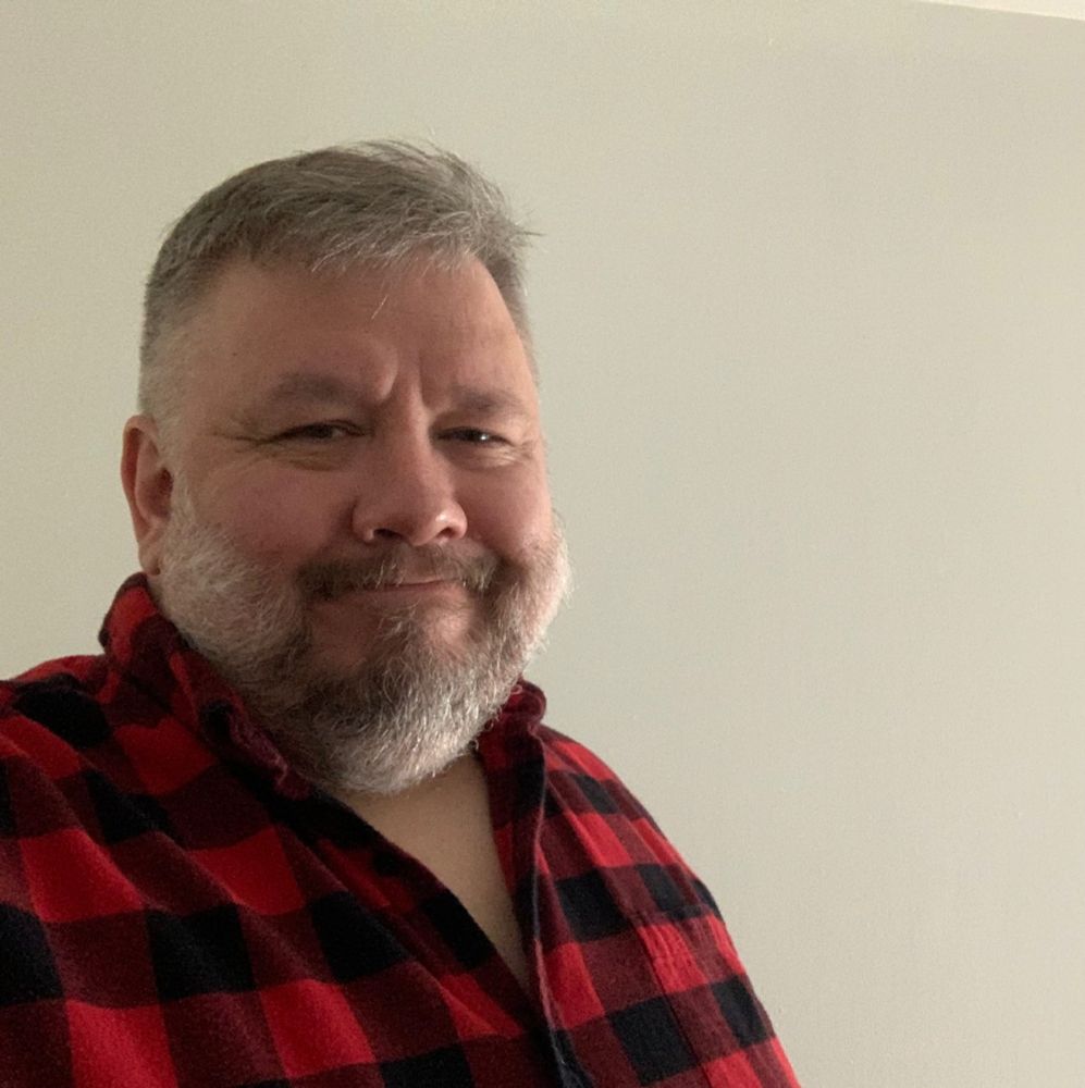 Chunky white guy with salt and pepper hair smiling in a red and black flannel shirt