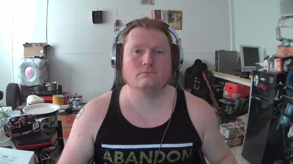 A sweaty, greasy ginger whose long red hair pulled back in a ponytail. A disheveled and cluttered office is seen in the background.