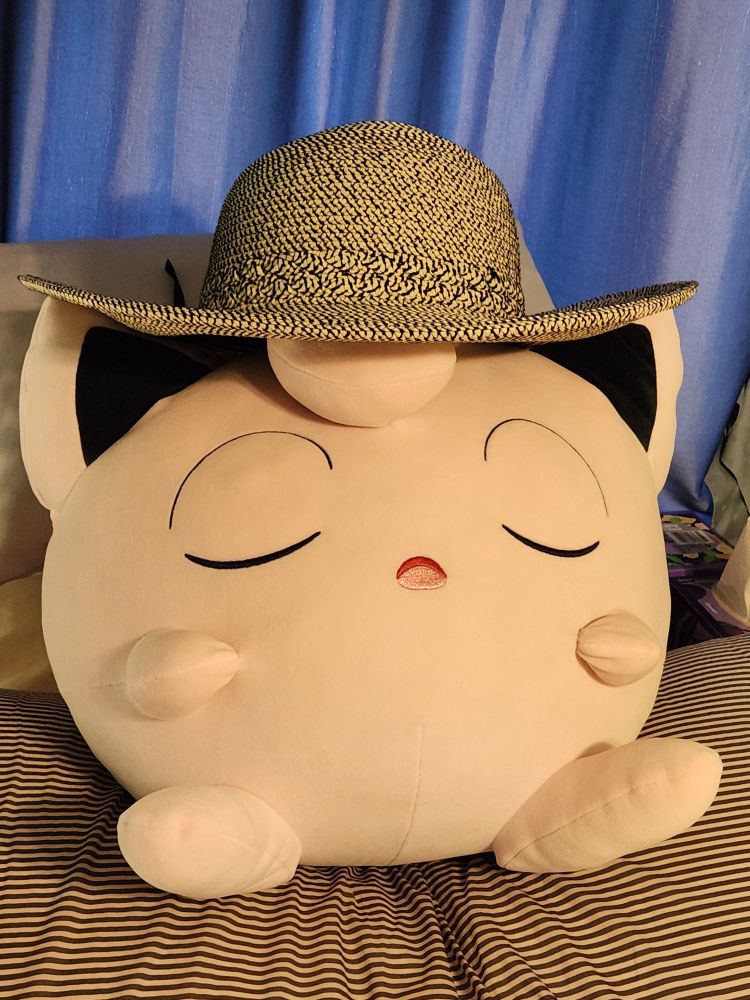 a plushie of the pokemon jigglypuff is sitting on a bed wearing a straw hat and has a sleepy expression on its face