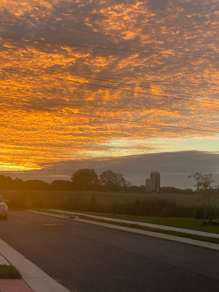 Clouds obscure the sun as it rises. In the distance is a cornfield with a farm silo.
