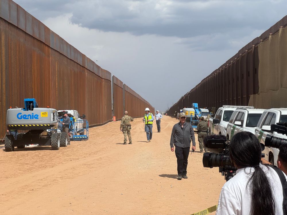 A dirt strip about the width of a two-lane highway between two border fences, with some cars, construction equipment, and personnel in view.