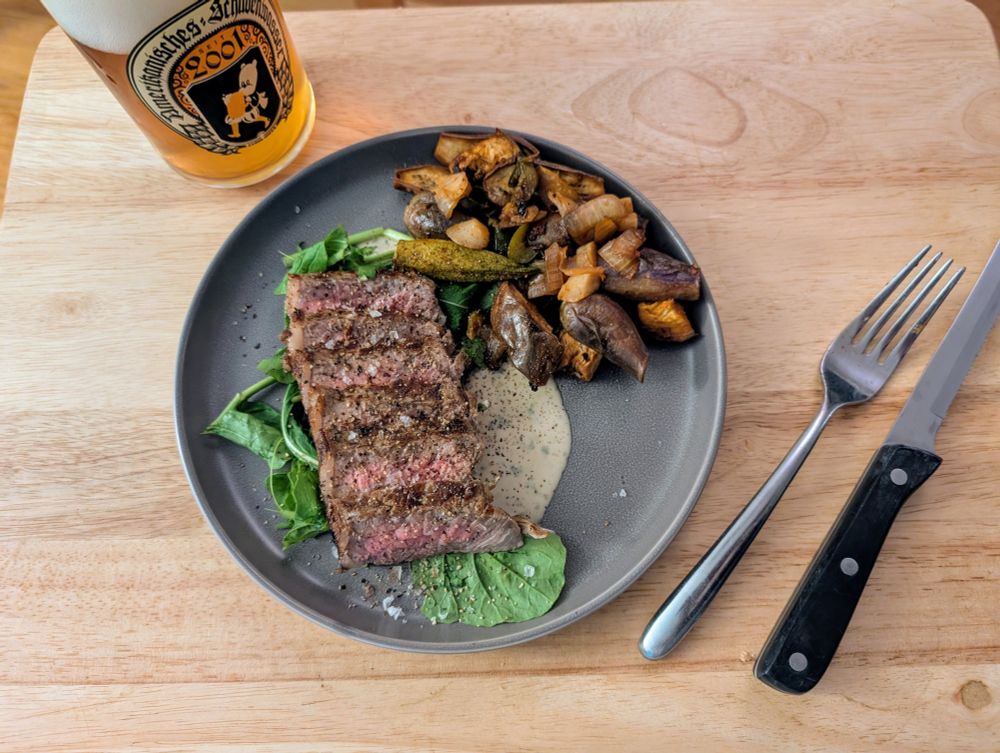 NY strip, arugula, tonnato, gochujang roasted eggplant/okra/jalapeno/alliums on a plate A glass of beer and utensils are nearby 