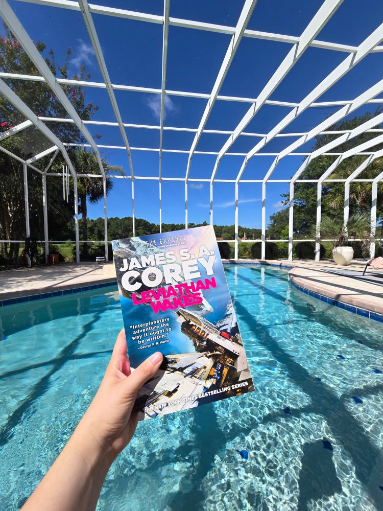 A photo of my hand holding a paperback copy of James SA Corey's Leviathan Wakes in front of a blue in ground pool that is covered by a patio screen. The sky is bright blue with a few whispy clouds in the sky and it is sunny. 