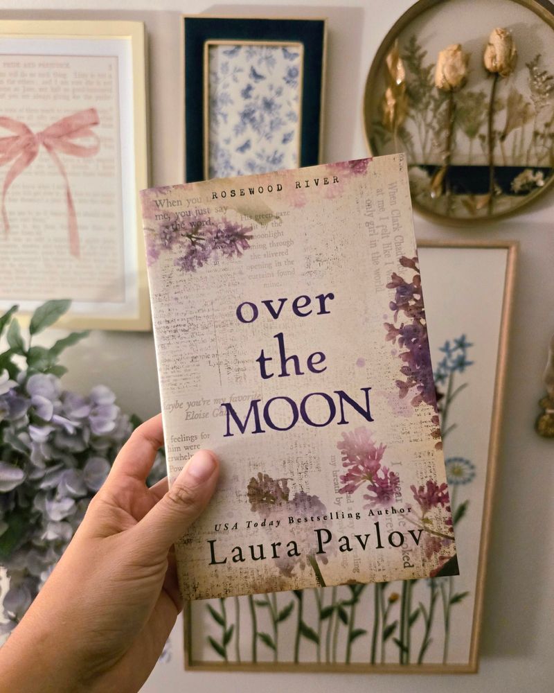 A photo of my hand holding up a paperback copy of Over the Moon by Laura Pavlov (the special edition copy with a lilac floral theme) in front of a wall collage of different floral items and photos in frames. 