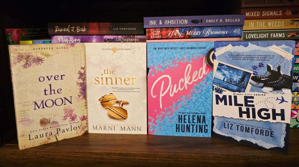 A photo of four paperbacks facing outward towards the camera. The paperbacks are sitting on a wooden shelf and behind the paper bags are other stacks of romance books. The paperbacks facing out words are all four from Entangled's Amara imprint. From left to right they are: Over the Moon by Laura Pavlov, The Sinner by Marni Mann, Pucked by Helena Hunting and Mile High by Liz Tomforde.