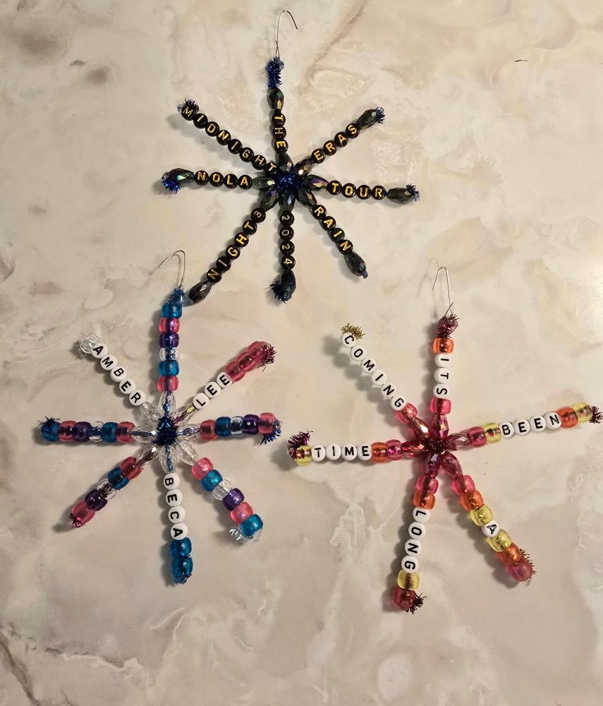 Chenille stem ornaments in the shape of snowflakes and Taylor Swift friendship bracelet themed. The top one is black and blue and navy themed and says The Eras Tour NOLA Night 3 2024 Midnight Rain. The bottom right is pink orange and yellow themed and says it's been a long time coming. The bottom right one is pink, blue, clear and purple themed and says Amber, Beca and Lee. 