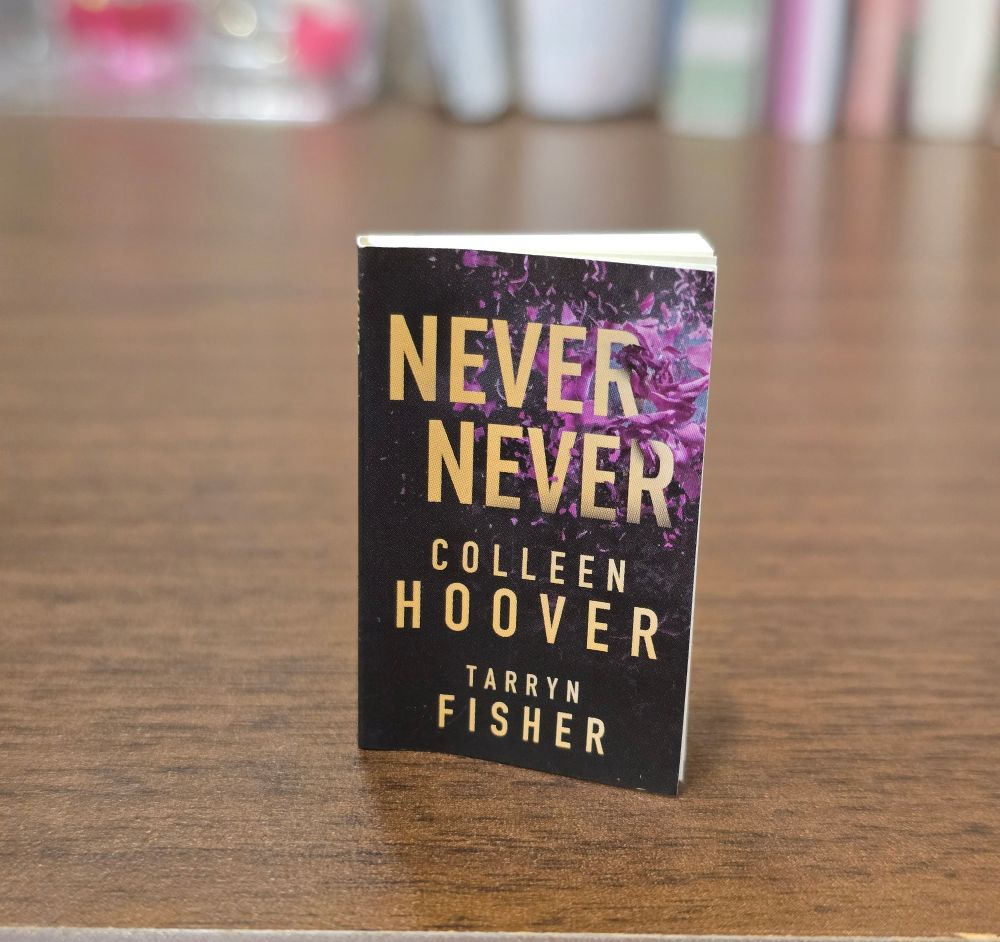 A mini book of Never Never by Colleen Hoover and Tarryn Fisher sitting on a wooden desk