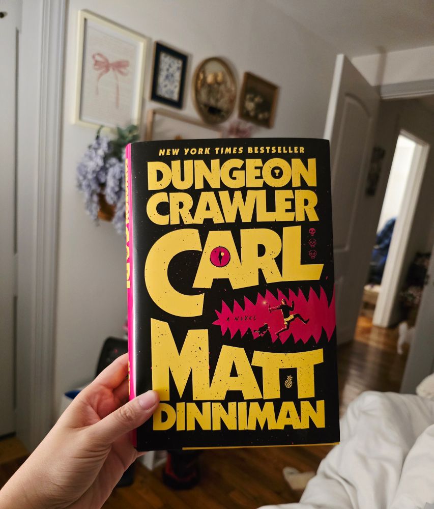 A photo of my hand holding up a hardcover copy of Dungeon Crawler Carl by Matt Dinniman. The background of the photo is my bedroom wall with an assortment of frames with flowers and bows on them
