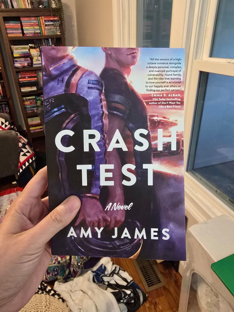 A photo of my hand holding a paperback copy of Crash Test by Amy James
