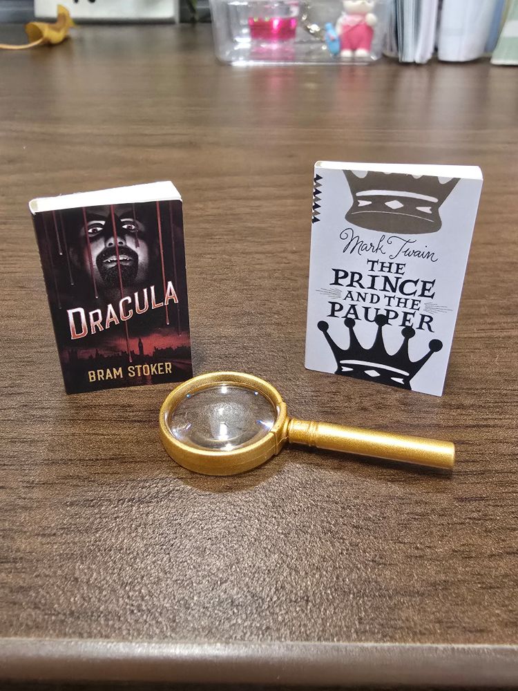 Two mini brand paperbacks (Dracula by Bram Stoker and The Prince and the Pauper by Mark Twain) along with a mini gold magnifying glass sit on a wooden desk