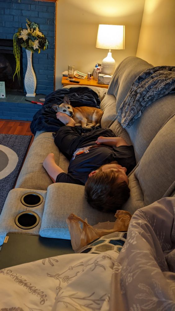 A photo of a guy sleeping on a couch with a brown furred dog resting their head on his foot. The guy is missing his hands and his right leg