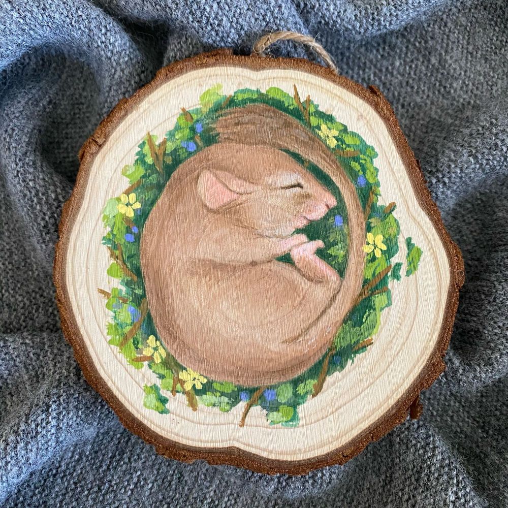 A brown mouse curled up in a circle asleep lays on a bed of green moss combined with twigs, yellow flowers, and blue berries. The scene is painted on a slice of wood with bark on the edges. Under the wood slice is a background of rumpled gray knitted material.