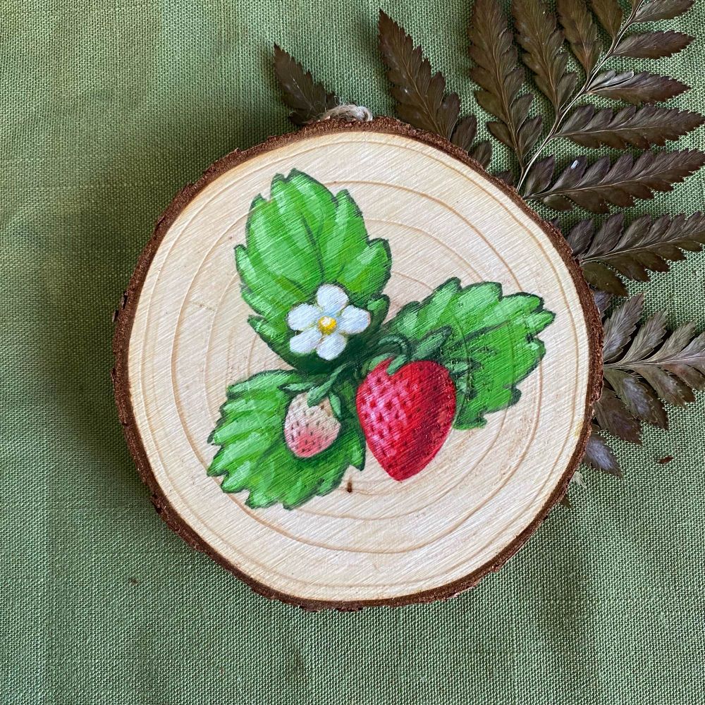Painting of one large strawberry, a small strawberry, and a white blossom surrounded by three green serrated leaves on a circular slice of wood. The slice of wood sits on a medium Olin green background with a fern leaf sticking out of the upper right.