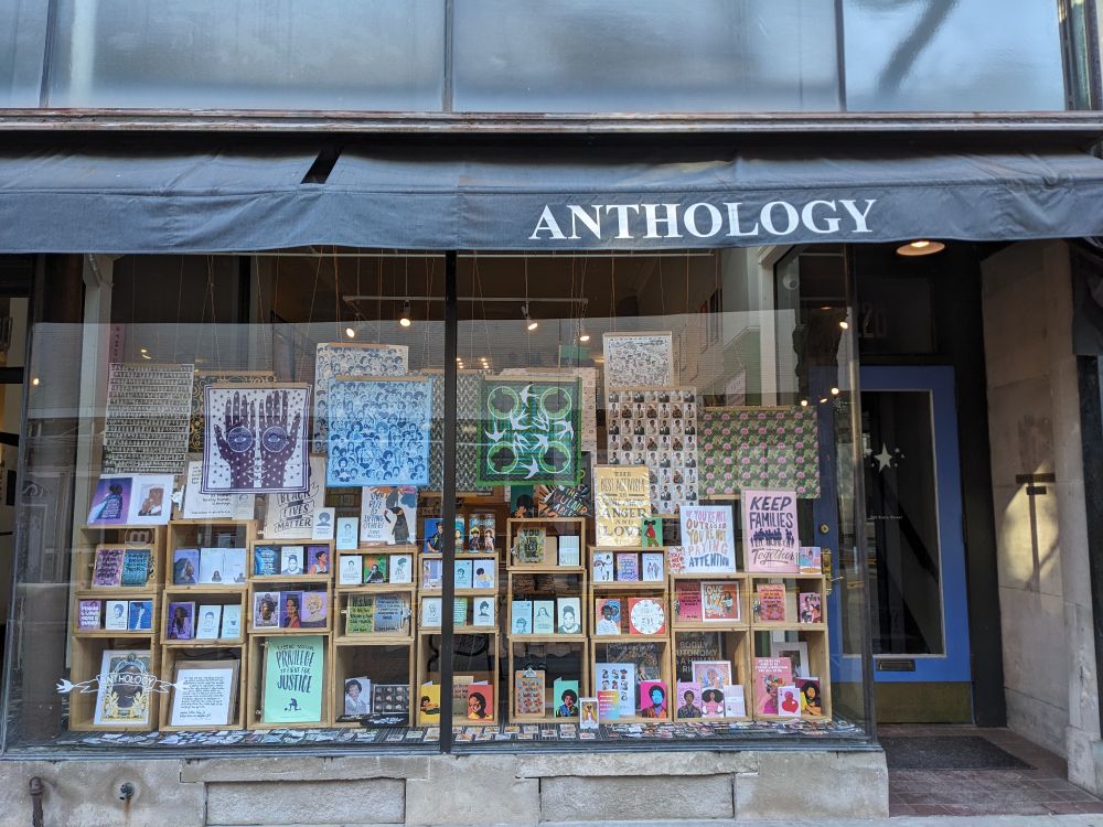 Store window with wooden cubbies holding cards and prints and other merchandise,  bandanas and sheets of wrapping paper hanging. Images of Black artists and poets and activists, "keep families together," "the best activism is equal parts anger and love," "if you're not outraged, you're not paying attention" posters.