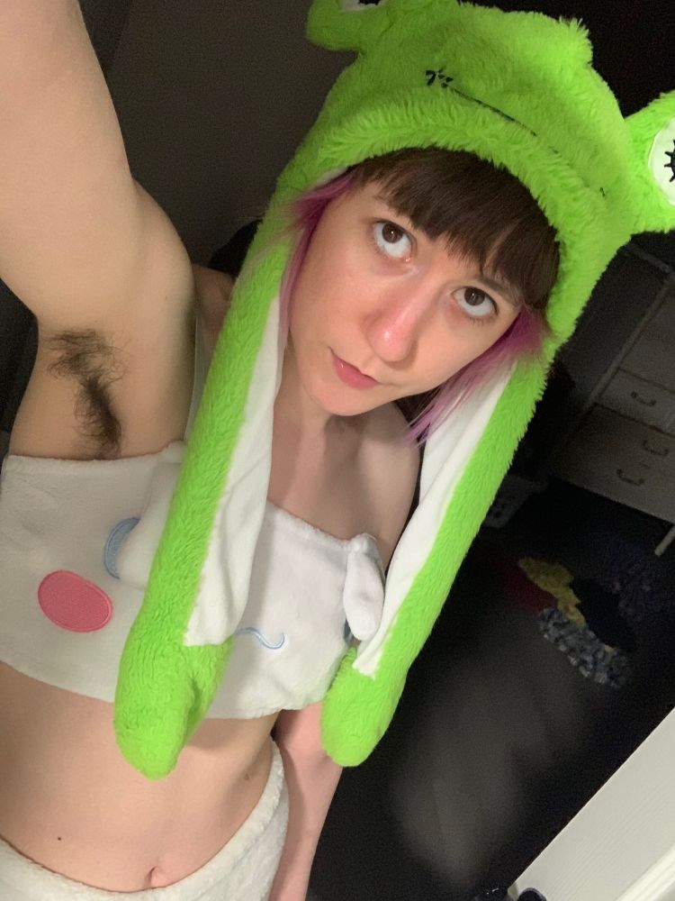 Charlie, a thin white femme person with short brown and purple hair stands in a doorway. The photo is selfie-style and you can see her armpit hair. She is wearing a white two piece fluffy lingerie set. The top is meant to look like the Sanrio character Cinnamaroll. She is wearing a fuzzy green frog hat.