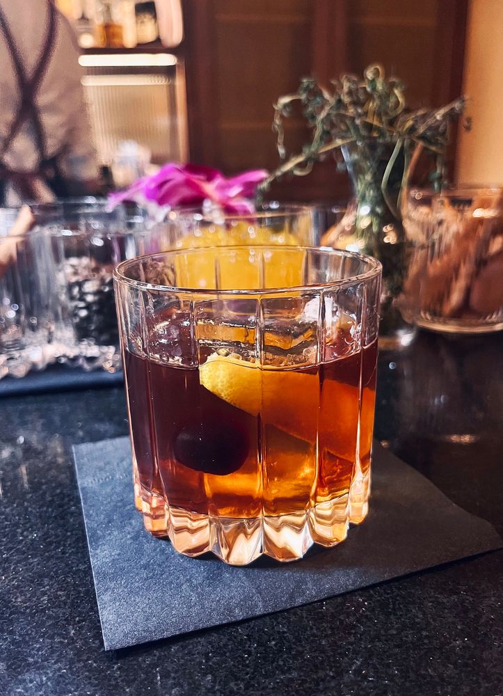 in a rocks glass on a black napkin on a black stone bar in a South American steakhouse, a reserve Guatemalan rum cocktail with a large, clear ice cube, simple syrup, angostura bitters garnished with orange peel and luxardo cherry