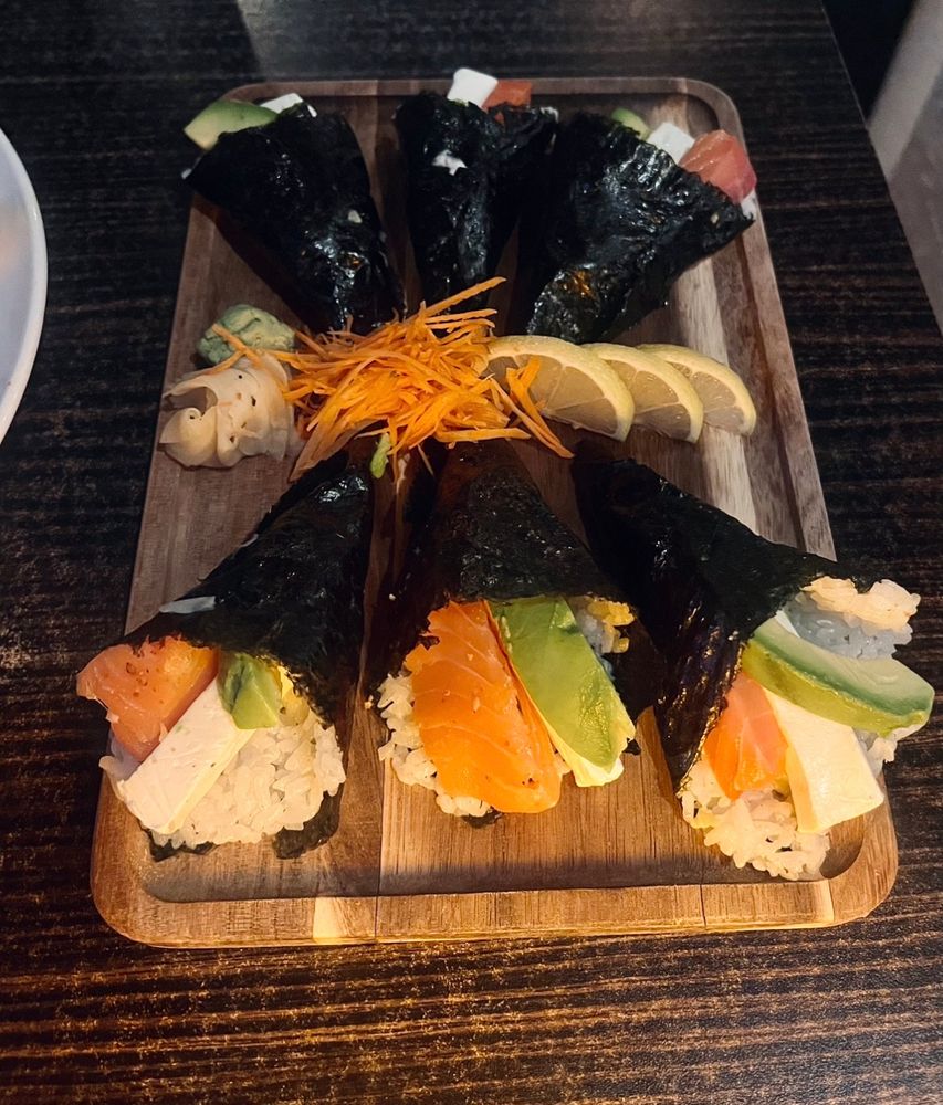 On a wooden board, six temaki filled with sushi rice and large slices of smoked salmon, cream cheese, and avocado garnished with wasabi, pickled ginger, and thinly sliced carrots 