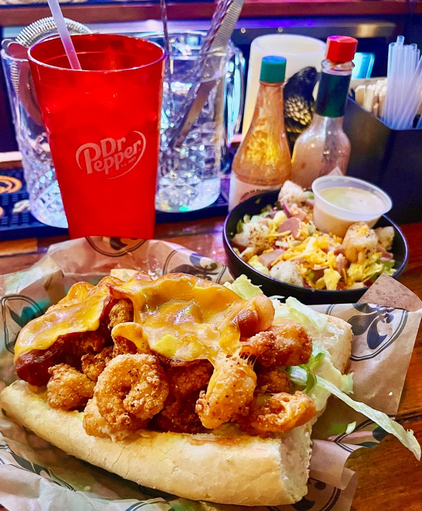 on the wooden bar of Mahony's Po-Boys & Seafood: a poboy on leidenheimer french bread dressed with lettuce, tomato, pickles, blue plate mayonnaise and cheddar cheese. in background, a chopped house salad (iceberg lettuce, tomato, shaved chisesi's vip ham, boiled egg, red onions, and creamy italian dressing), bottles of Tabasco and Crystal hot sauces, and a tall red Dr Pepper glass.