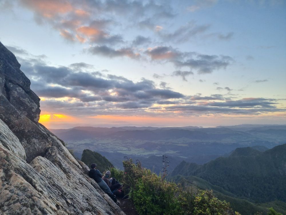 Photograph of a cloudy sky above a green landscape with a freshly risen sun creating an orange glow to the left of the sky. 