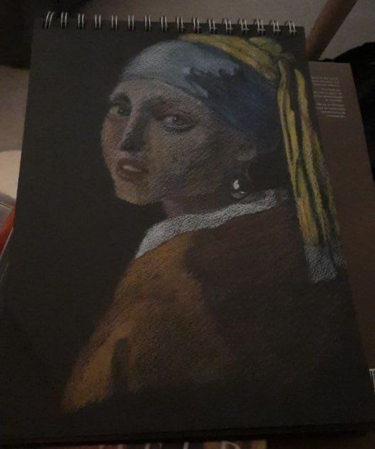 Drawing of The Girl with the Pearl Earring, in coloured pencils on a black sketchpad. The girl is turning over her left shoulder to look directly at the viewer. She has a light blue material headband, with yellow flowing material from her head scarf.
She has a rusty brown shawl with a white collar