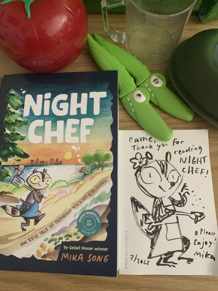 graphic novel "Night Chef An Epic Tale of Friendship with a Side of Deliciousness! (A Graphic Novel)
By Mika Song with racoon in overalls and a baby crow walking out on an adventure. Book is on table with cooking tools like funny alligator shaped paring knives and tomato and avocado shaped food storage. A doodle of main character: the cook racoon is also on table.