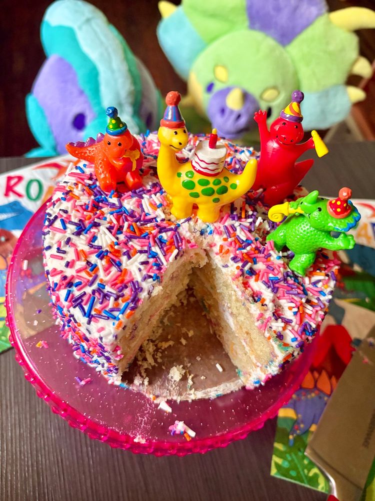photo of a confetti sprinkle cake with four colorful partying dino figurines on top. the cake is on a pink cake stand, on a black table, with dinosaur themed napkins. there are two Rawrcade plushies on the ground, positioned to look up at the cake. a slice has been taken out of the cake, a knife is laying on the napkins. 
