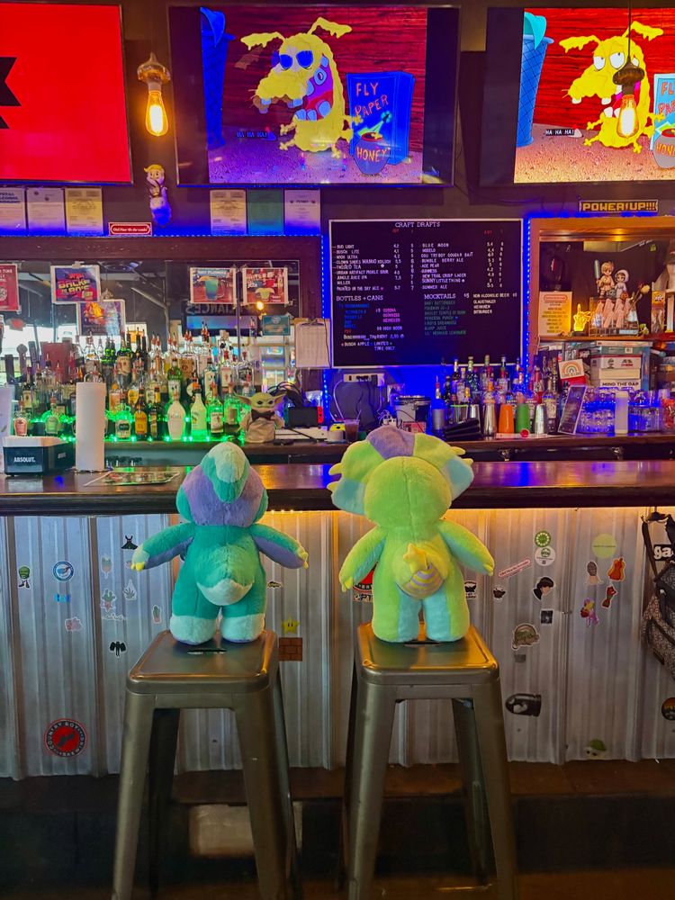 A photo of two Rawrcade dinosaur plushies sitting at a barcade, on stools. Courage the Cowardly dog is on t.v.