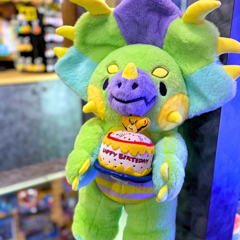 photo of a Rawrcade character Cade plush at an arcade with a prize counter in the background. Cade is holding a Build-A-Bear plush birthday cake prop.