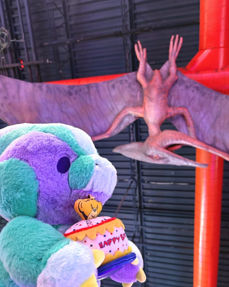 Photo of Rawrcade plush Fizz with a Build-a-Bear birthday cake prop, a pteranodon is in the distance. 