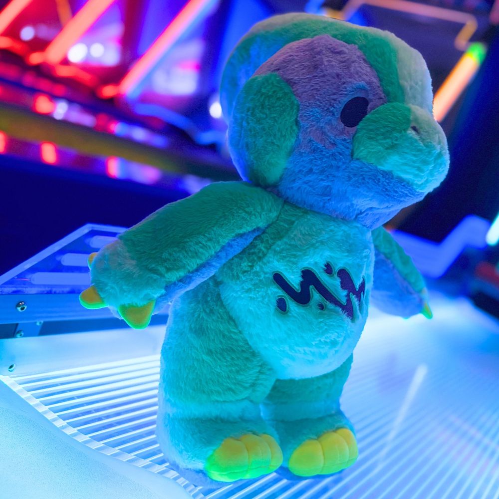 photo of a Rawrcade character Fizz plush at an arcade with neon lights in the background. 