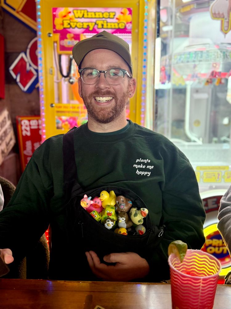 A photo of me sitting at a table wearing a green sweatshirt that says “plants make me happy” I have a green hat on and a black cross body bag. The bag is filled with rubber ducks in a variety of colors and designs. 