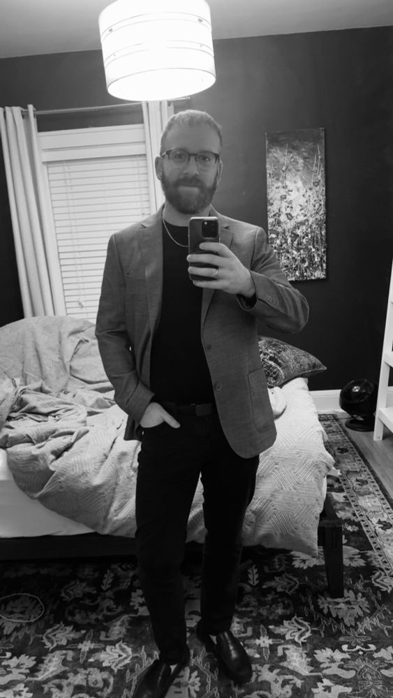 A monochrome photo of me in black pants, black loafers, thin black wool sweater and and burgundy sport coat that obviously shows up as grey because monochrome. I’m subtly smiling, my beard is looking full, and my hair is styled down 