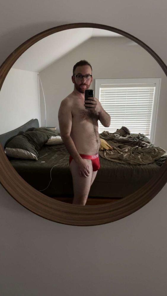 A mirror selfie of myself wearing red bikini style briefs. A messy unmade bed is in the background. 