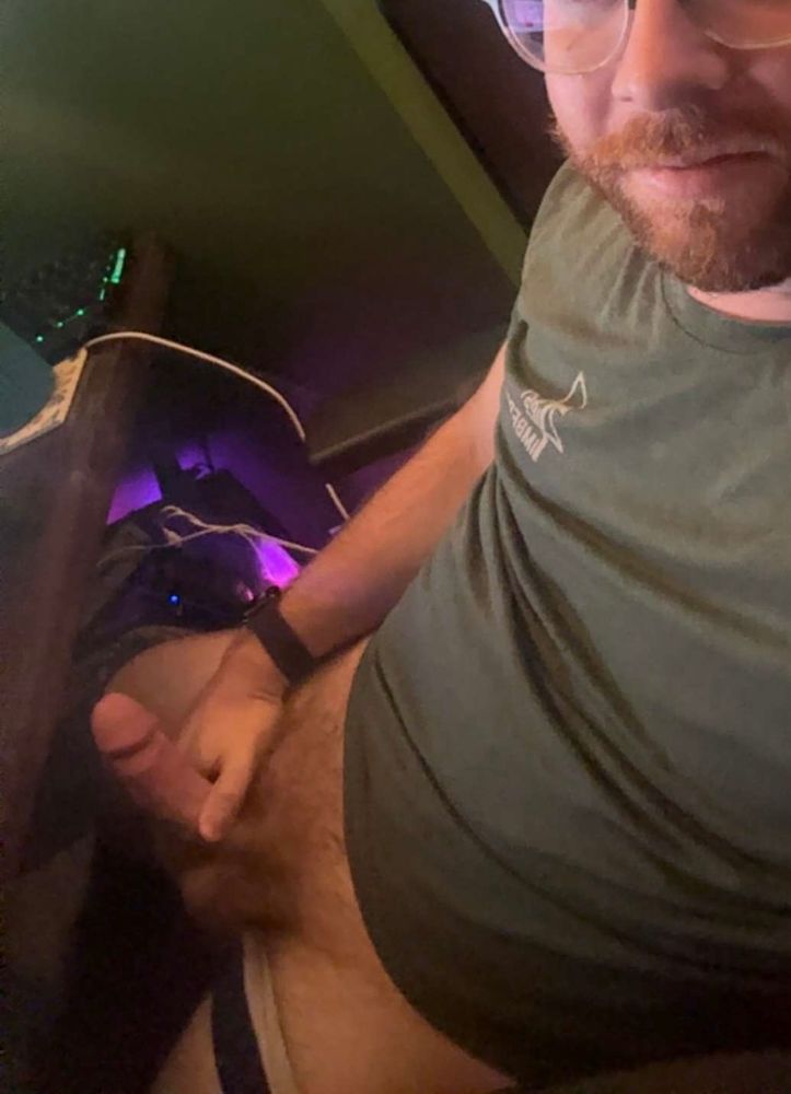 A photo of a man (me) sitting at a desk. His pants are off and his underwear are pulled down on his thighs. He’s holding his hard cock for the camera and wearing a green tshirt. 