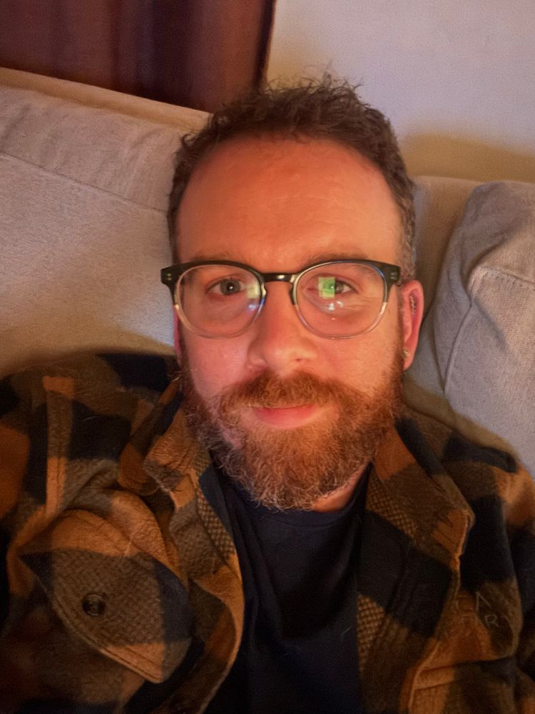 A selfie of me on the couch with a tan and blue checkered fleece shirt on and a pretty full beard with brown, red, and grey hair. 