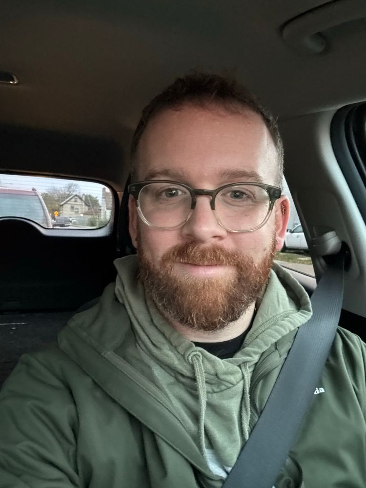 A selfie of me, a 30-something man with brown hair and a ginger beard in my car. I’m wearing glasses and a green sweatshirt with a green jacket on top of it. 