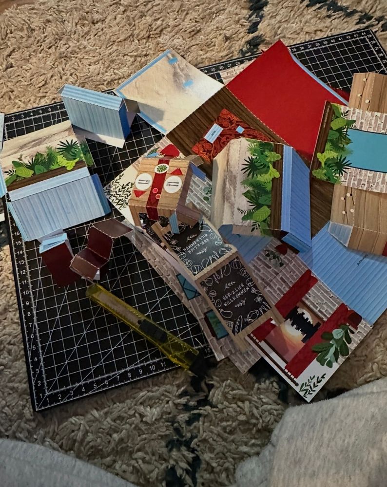 Auf dem Bild liegen mehrere bunte, ausgeschnittene Papierstücke auf einer schwarzen Schneidematte mit weißem Raster. Neben den Papierstücken befindet sich ein gelbes Schneidemesser. Die Matte liegt auf einem beigen Teppich mit flauschiger Struktur. Die Papierstücke zeigen verschiedene Muster, darunter Holzoptik, Pflanzenmotive, Backsteinmuster, Schriftzüge und farbige Flächen in Rot, Blau und Weiß. Einige Teile sind bereits gefaltet.