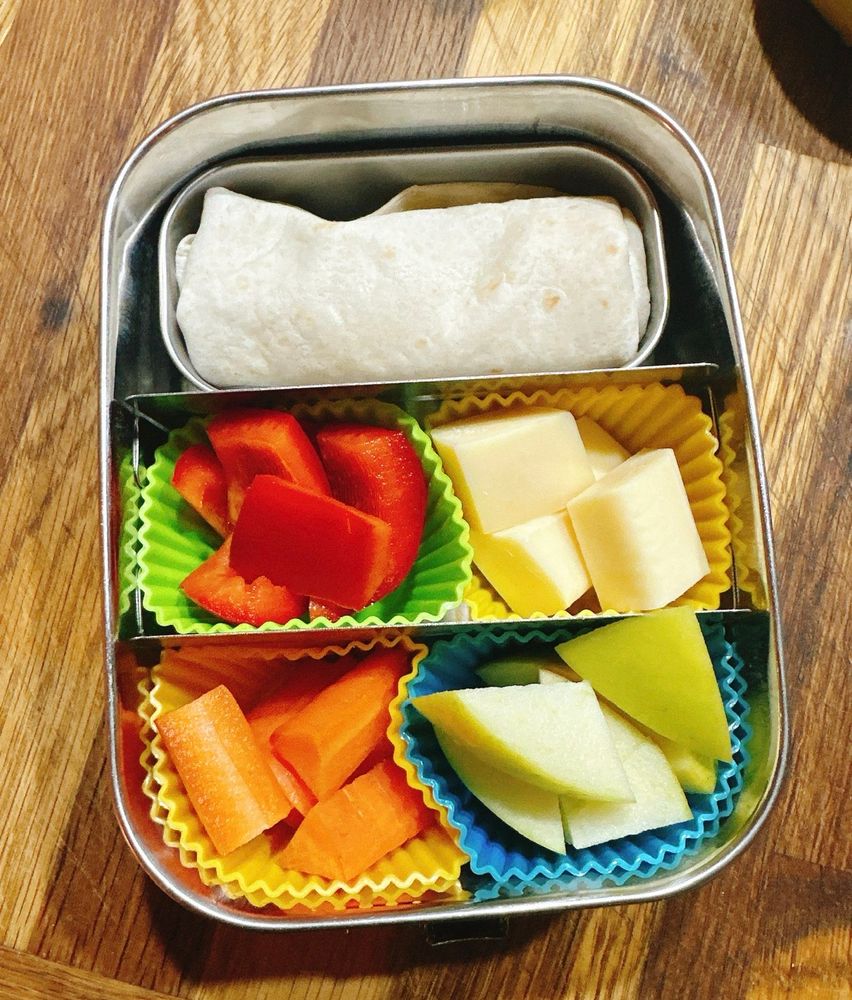 Auf dem Bild ist eine geöffnete Metall-Brotdose zu sehen. In der oberen Hälfte liegt ein kleiner, zusammengerollter Wrap. Darunter ist die Box durch zwei Trennwände in drei Fächer geteilt. Links unten liegen Stücke einer roten Paprika in einer grünen Silikonform. Rechts daneben liegen helle Käsewürfel in einer gelben Silikonform. Links unten liegen geschnittene Möhrenstücke in einer gelben Silikonform. Rechts unten liegen helle Apfelstücke in einer blauen Silikonform. Die Brotdose steht auf einer Holzoberfläche.
