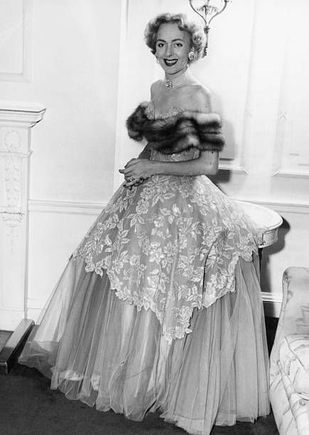 Jorgensen wears an off-the-shoulder ball gown with a fur-trimmed neckline, lace overlay, and voluminous chiffon skirt with a choker necklace, large earrings, and glam makeup.
