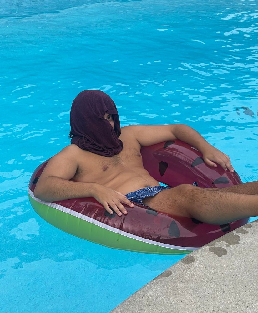 a photo of me sitting in a watermelon themed inflatable tube in a pool. I’m shirtless and wearing a blue bathing suit with a dark red shirt wrapped around my head to keep the sun out of my face 